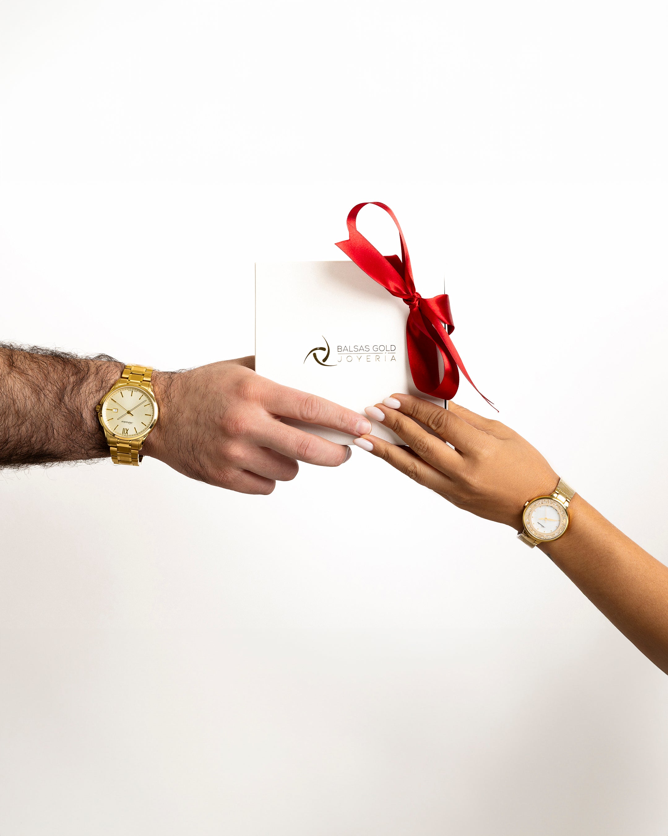 Manos entregando una caja de regalo de Balsas Gold Joyería con un moño rojo, usando relojes de oro. Imagen de campaña navideña 2025.