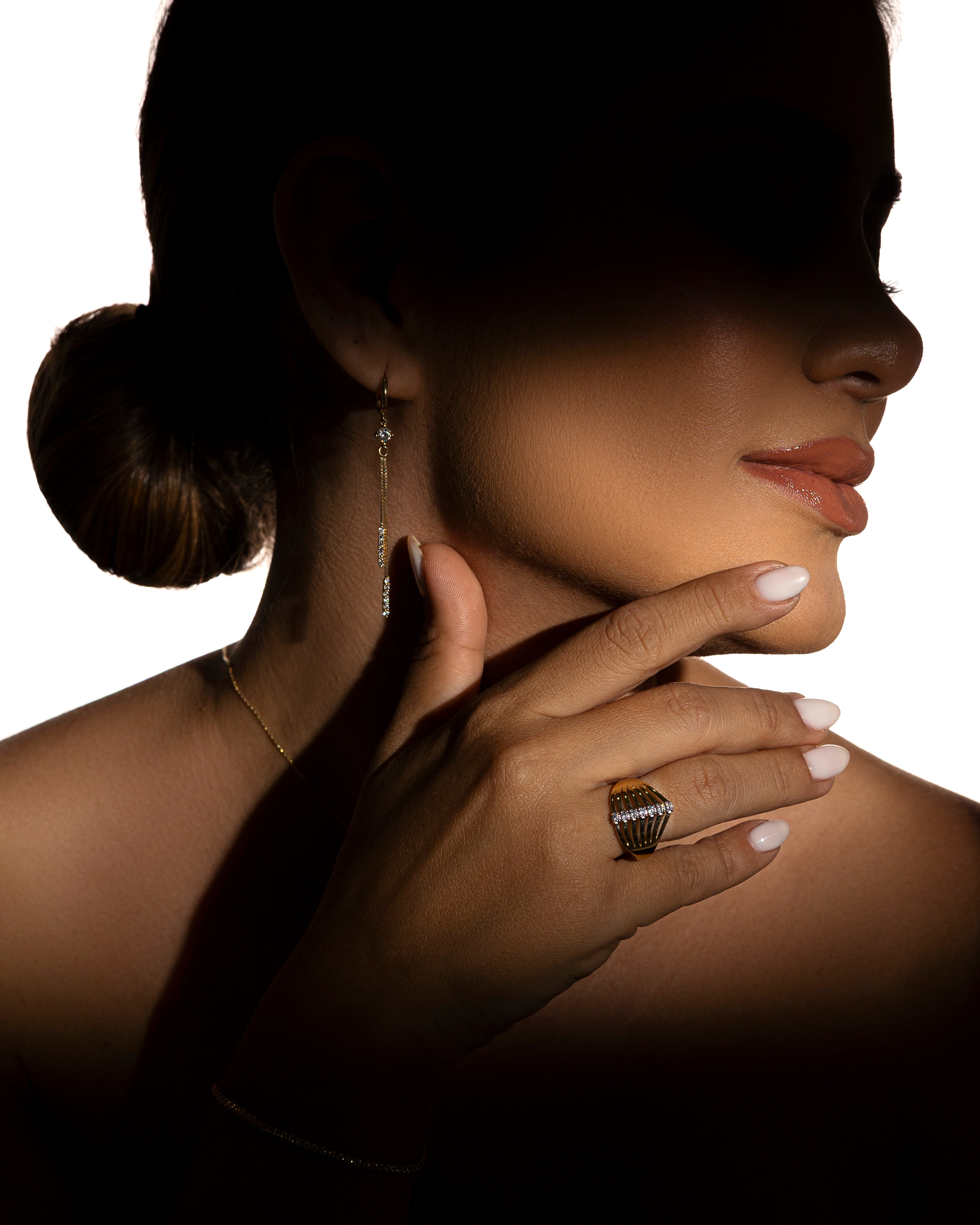 Primer plano del rostro y mano de una mujer usando joyería de oro: anillo, arete largo y collar delicado, con iluminación dramática.